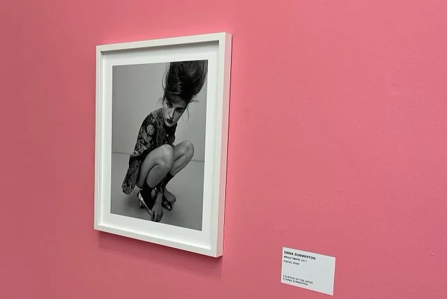 Black-and-white fashion photograph of a crouching woman with a beehive hairstyle, framed and displayed on a pink wall in an art gallery.