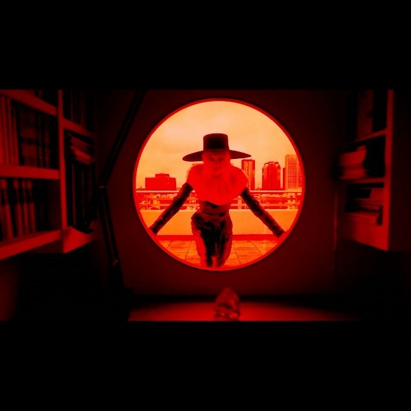 A person wearing a hat stands in a round doorway overlooking a city skyline. Red lighting casts a dramatic mood, with bookshelves visible.