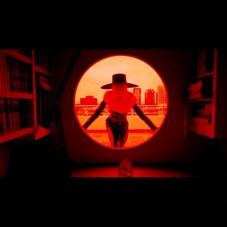 A person wearing a hat stands in a round doorway overlooking a city skyline. Red lighting casts a dramatic mood, with bookshelves visible.