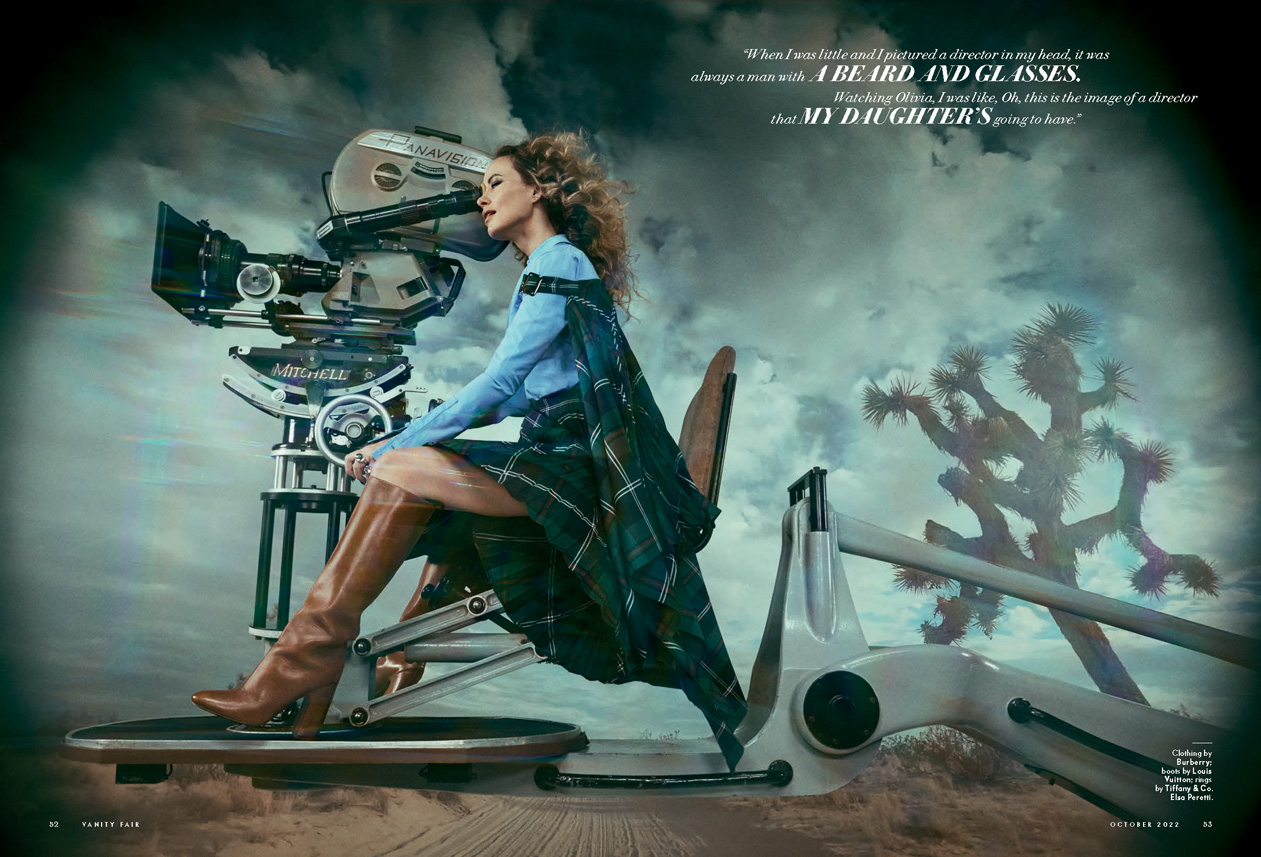 Adult woman with curly hair sitting on film-camera crane near a vintage motion-picture camera, wearing a plaid skirt and brown boots.