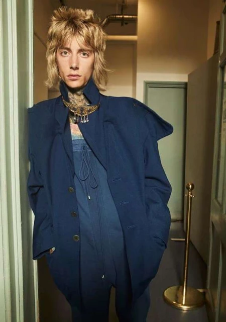 A person with blond hair and a large blue blazer is standing indoors, looking at the camera with neutral facial expression.