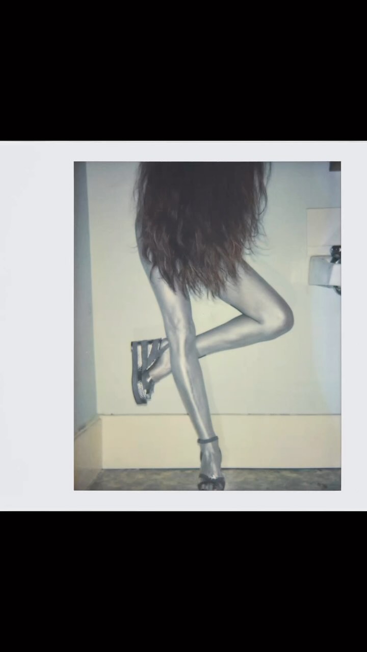 Abstract shot of a person wearing shiny silver leggings and high heels, with long dark hair, posing in front of a white wall.