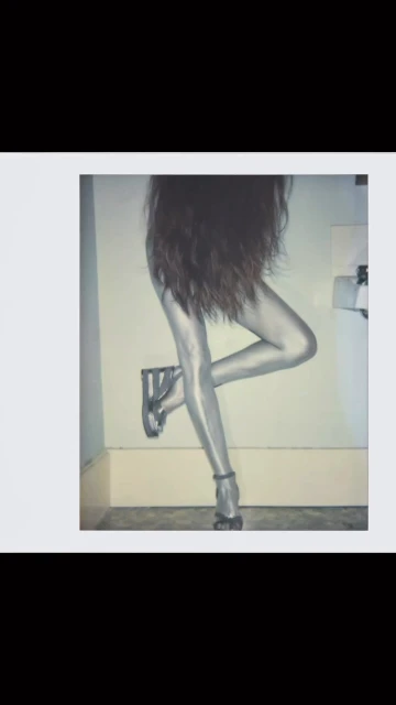 Abstract shot of a person wearing shiny silver leggings and high heels, with long dark hair, posing in front of a white wall.