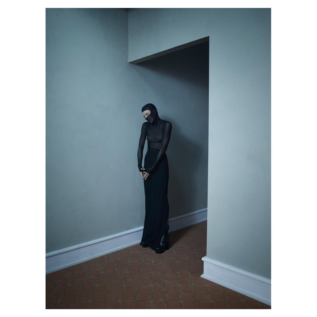 A person stands in an interior wearing a black, semi-sheer turtleneck top, black maxi skirt, and a head covering in a doorway.
