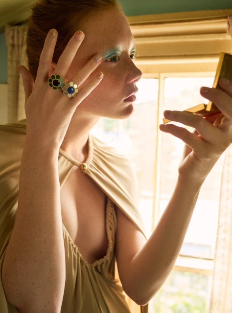 Woman with freckles looking in compact mirror, wearing a beige gown and flower rings, with window light coming from behind.