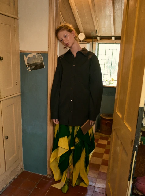 A tall, red-haired woman in an oversized dark shirt and bright yellow and green floral skirt stands in a doorway inside a home.