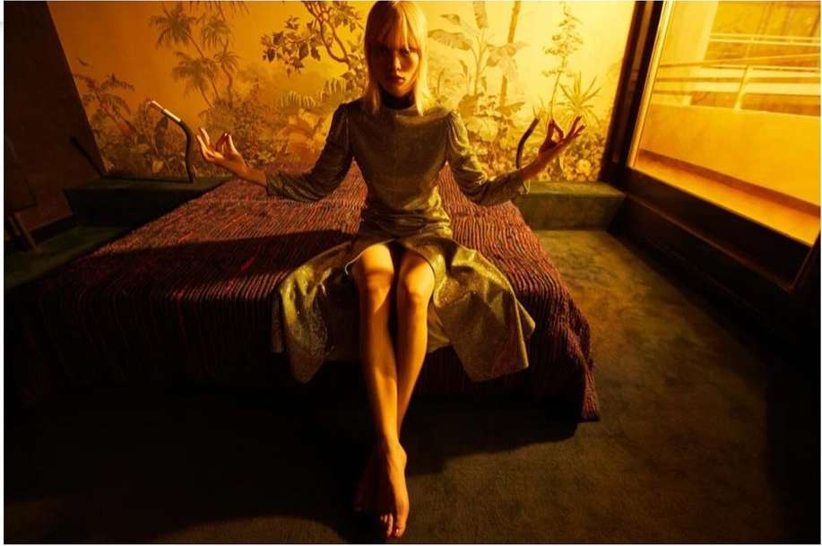 Fashion shot of a slender blonde woman sitting on a bed with legs crossed, arms raised, and hands in a symbolic mudra gesture.