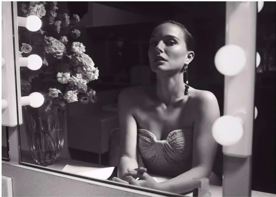 Monochrome portrait of an adult female sitting in front of a lighted vanity mirror with a vase of roses next to her.