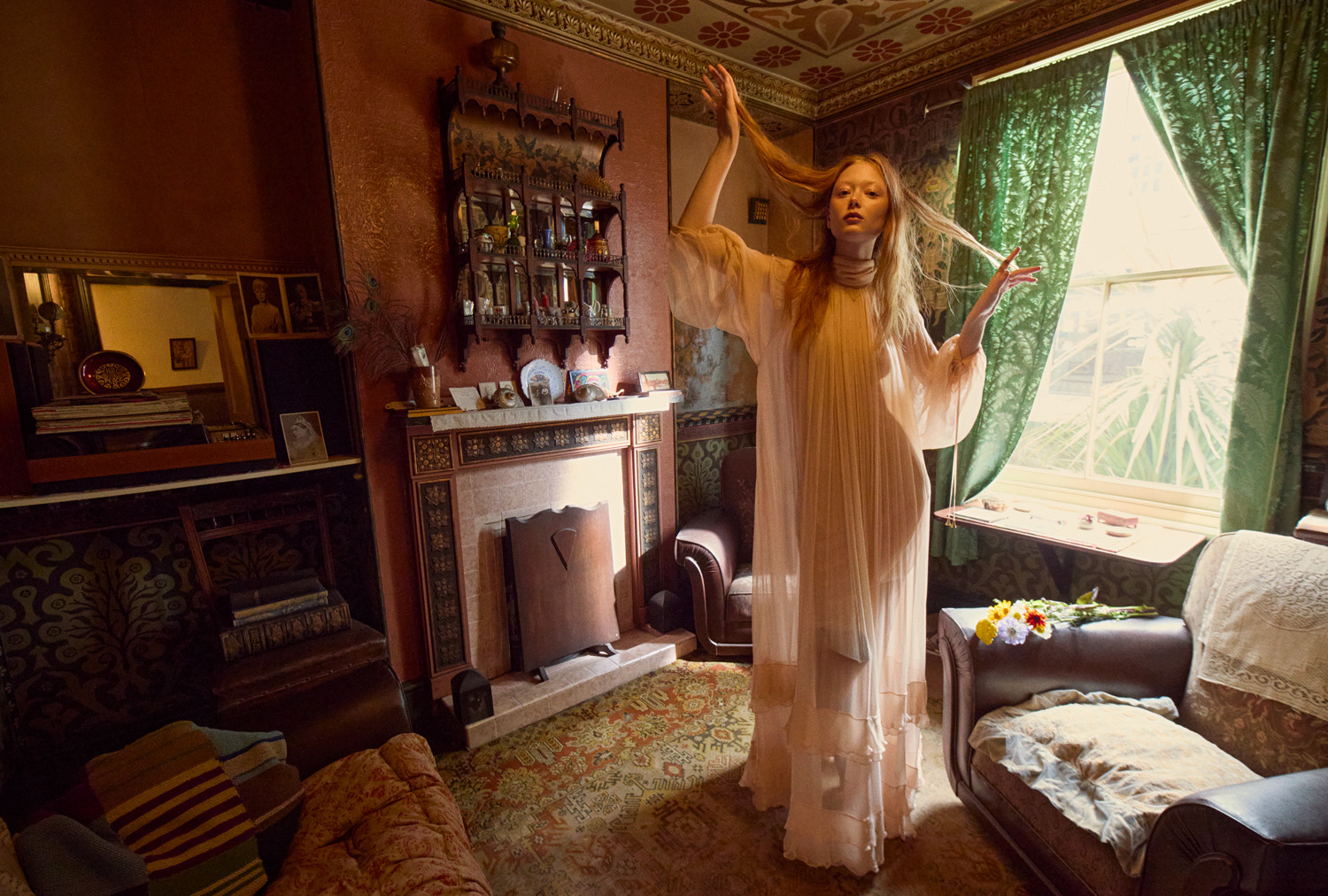 A fair-skinned woman in a flowing white gown stands in a sunlit, patterned interior with vintage furniture and green drapery, in a fashion editorial.