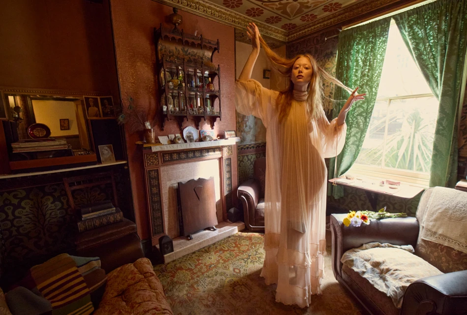 A fair-skinned woman in a flowing white gown stands in a sunlit, patterned interior with vintage furniture and green drapery, in a fashion editorial.