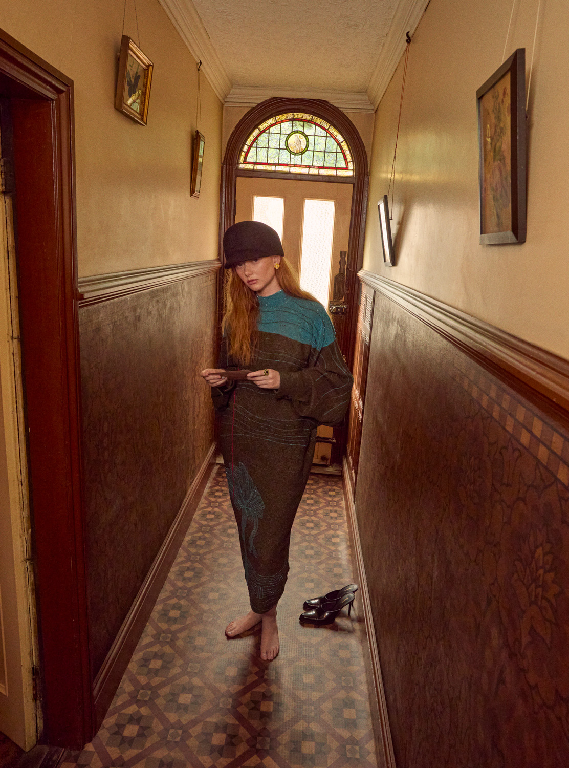 A woman in a long patterned dress and cap stands in a hallway with paintings and dark wood trim, holding a small object.