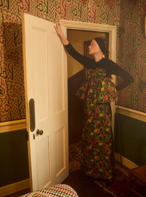 A model poses in a floral dress while opening a white door in a vintage-style room with patterned wallpaper and antique furniture.