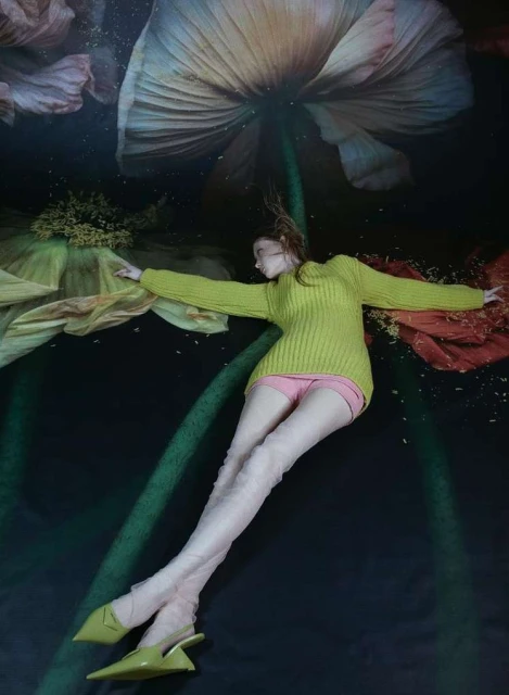 A female fashion model in a yellow sweater and pink shorts lies on a dark surface with large painted flowers in a studio setting.