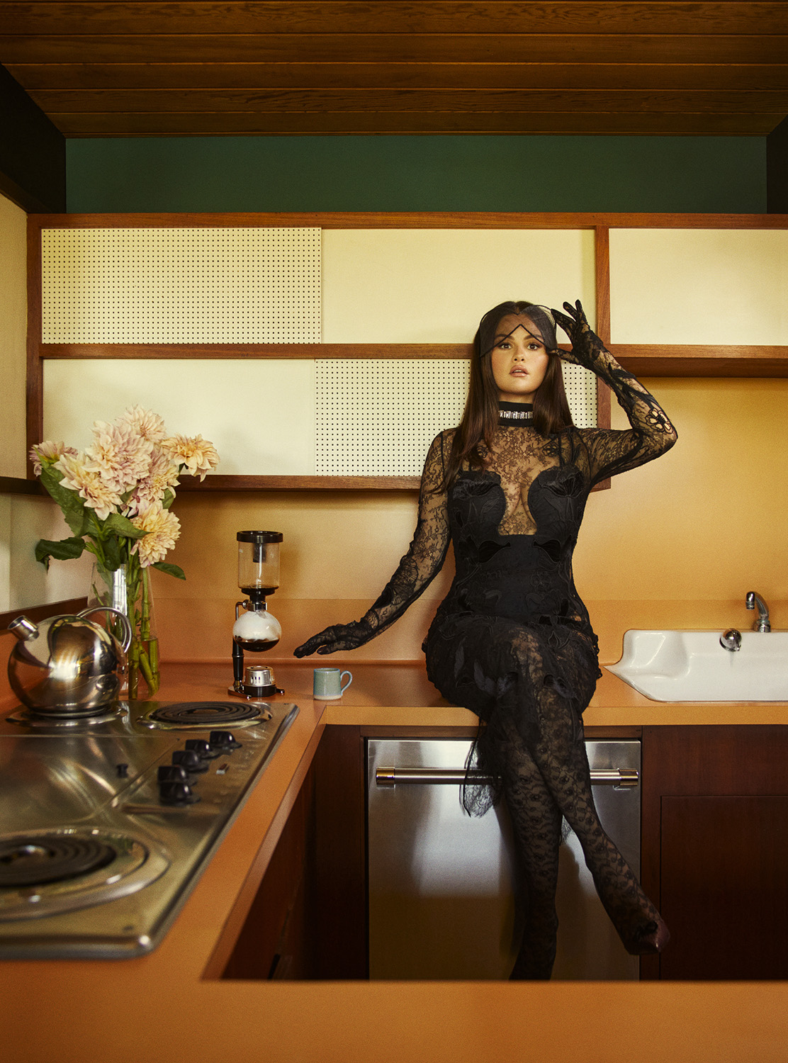 Woman in a black lace dress and face covering poses on a kitchen counter next to flowers, a coffee maker, and a sink.