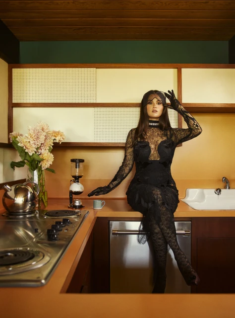 Woman in a black lace dress and face covering poses on a kitchen counter next to flowers, a coffee maker, and a sink.
