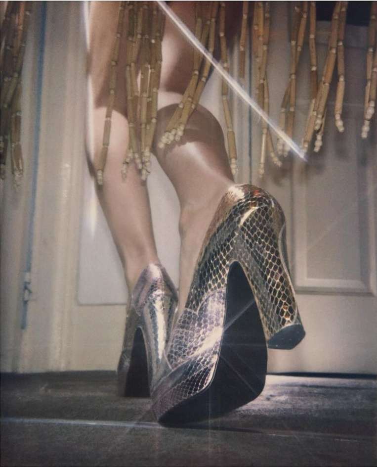Close-up of a woman's legs and shiny, snakeskin-patterned high heels on a gray floor with gold fringe in the background.