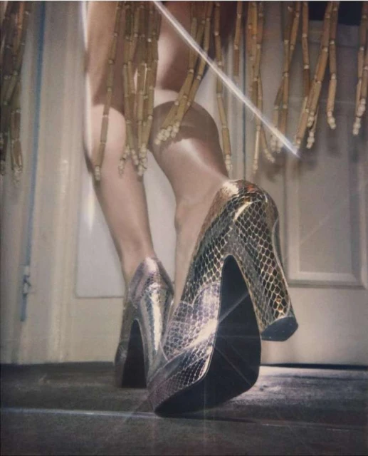 Close-up of a woman's legs and shiny, snakeskin-patterned high heels on a gray floor with gold fringe in the background.