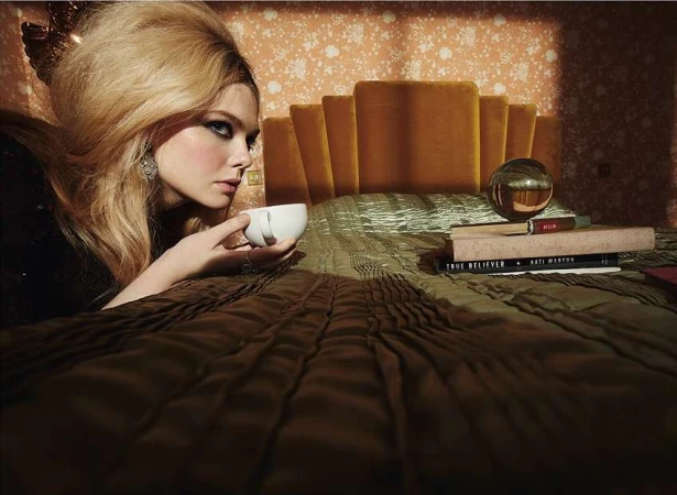 Blonde woman with voluminous retro hair holding a white teacup while lying on a bed with a velvet headboard by artist Emma Summerton