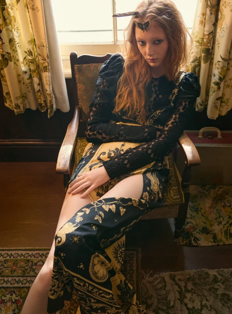 Elegant red-haired woman in patterned black and gold gown sitting in chair with antique floral wall in the background in moody indoor setting.