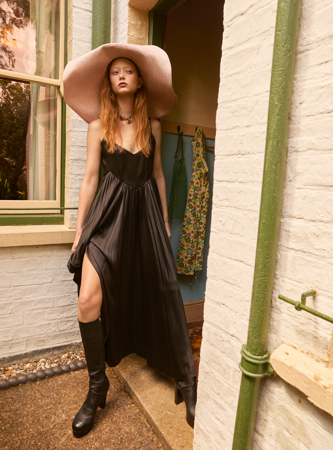 A fair-skinned woman poses in a black dress and oversized hat, her knee-high boots adding to the fashion statement in an outdoor setting.