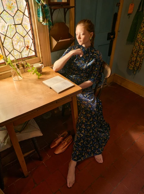 Red-haired woman in floral dress seated at wooden table near window with stained glass, reading a book in a rustic interior.