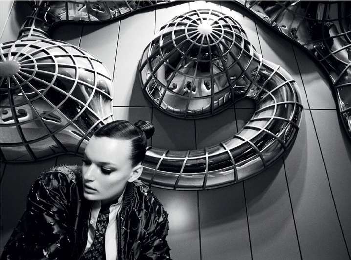 Fashion portrait of a woman in a patterned jacket with metallic sphere sculptures against a grey tiled wall, photographed indoors.