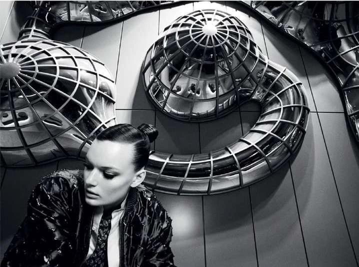 Fashion portrait of a woman in a patterned jacket with metallic sphere sculptures against a grey tiled wall, photographed indoors.