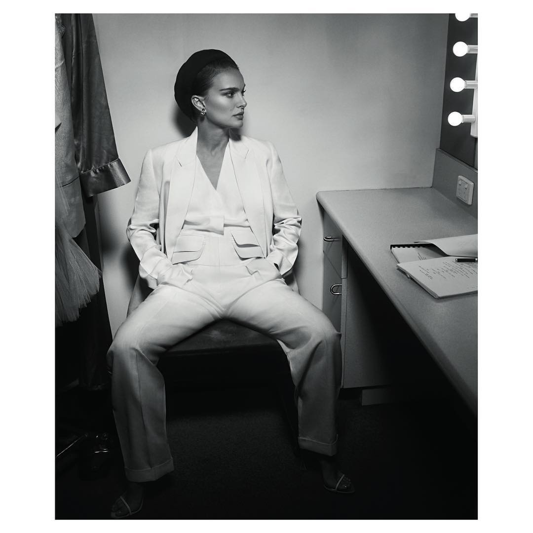 Black and white shot of a woman in a white suit, sitting in a dressing room with a mirror and clothing rack in the background.