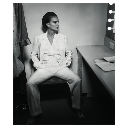 Black and white shot of a woman in a white suit, sitting in a dressing room with a mirror and clothing rack in the background.