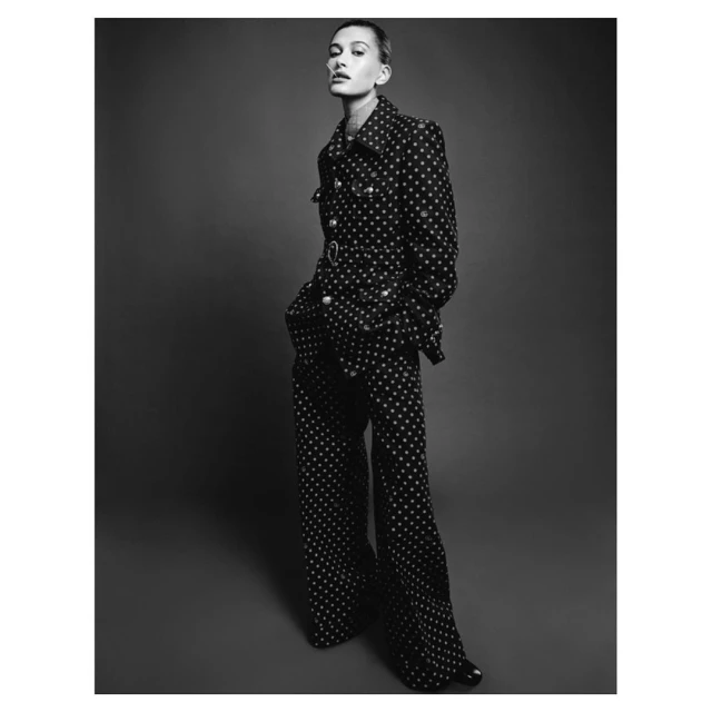 Black and white portrait of a female model posing in a patterned jacket and wide-leg pants against a dark backdrop in a studio setting.