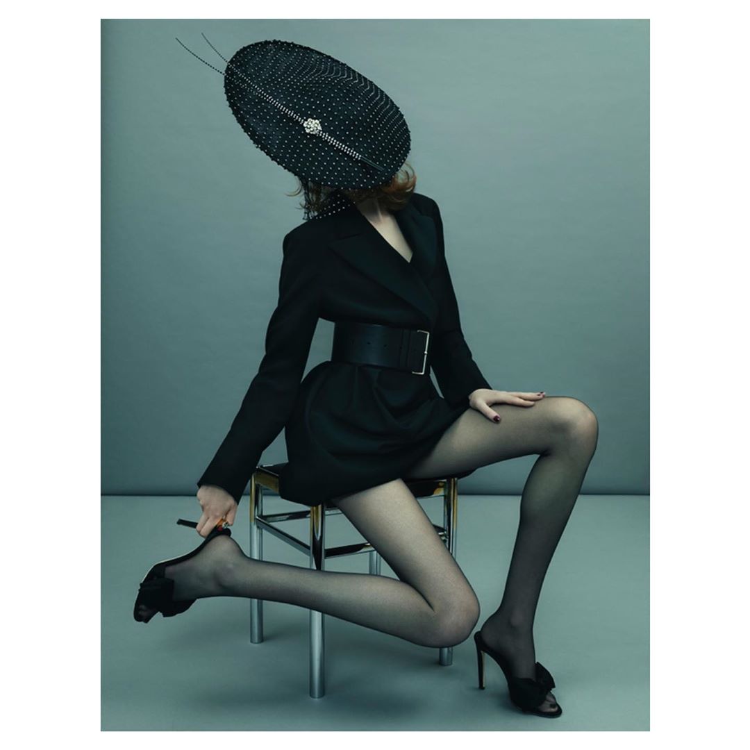 Woman in a black jacket, belt, and sheer black stockings sitting on a chrome chair with a large dotted black hat indoors.