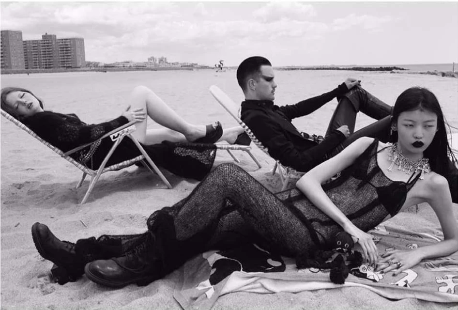 Three fashion models pose on a sandy beach wearing dark clothing, showcasing editorial style, with an urban building in the background.