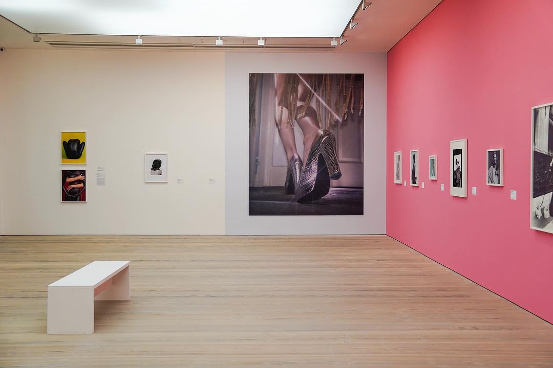 Bright art gallery interior features a large photograph of sequined heels with additional framed photos on a pink wall, and a white bench.