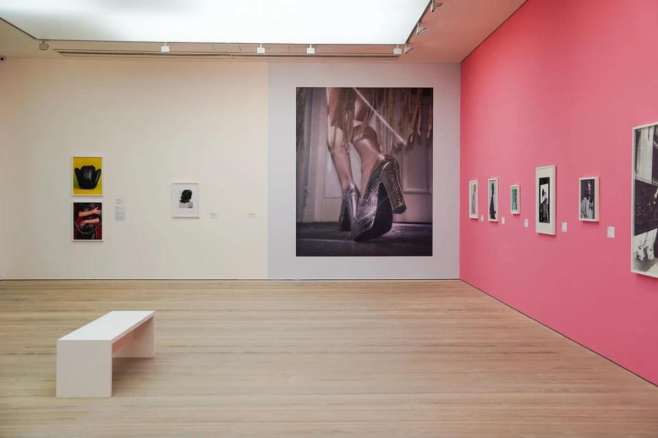 Bright art gallery interior features a large photograph of sequined heels with additional framed photos on a pink wall, and a white bench.