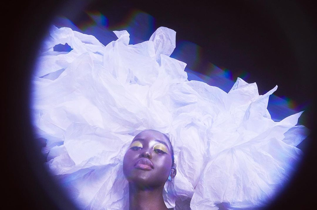 Artistic portrait of a young Black woman with gold makeup and an elaborate white headpiece, set against a dark background with soft lighting.