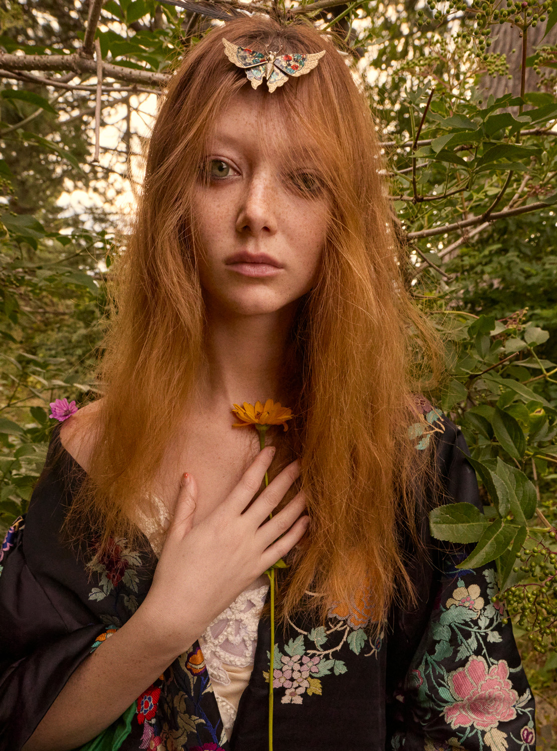 A woman with red hair and a floral robe holds a yellow flower against a backdrop of green foliage and dappled sunlight, creating a serene portrait.