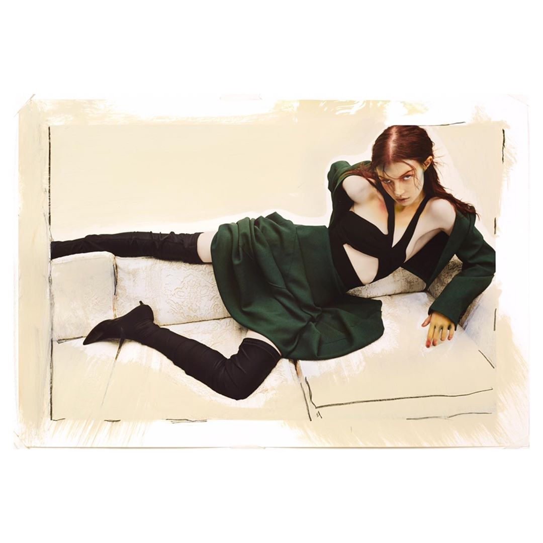 Fashion editorial shot of a reclining adult female wearing a black top, green skirt, jacket, and high black boots on a white couch.