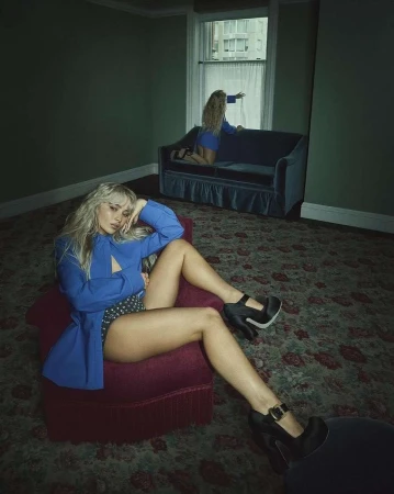 Fashion shot of a blonde woman in a blue blazer and polka dot shorts sitting on a red chair, with a second woman on a couch near a window.