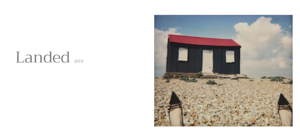 Point of view shot of black high heeled shoes on a pebble beach facing a small black corrugated building with a red roof under a blue sky.