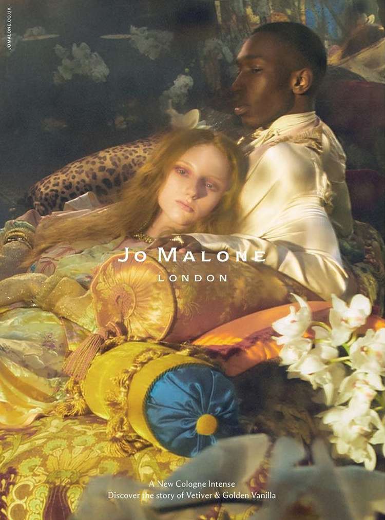 An interracial couple poses amid rich textiles and flowers in a perfume advertisement for Jo Malone London, with soft lighting.
