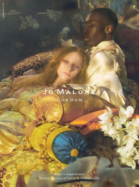 An interracial couple poses amid rich textiles and flowers in a perfume advertisement for Jo Malone London, with soft lighting.