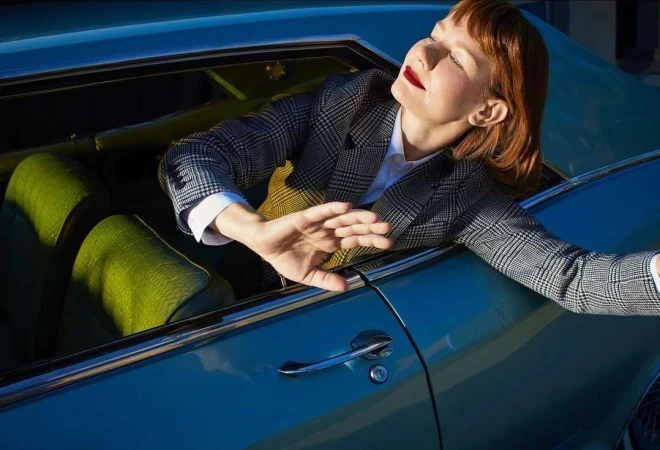Red-haired woman in houndstooth blazer leaning out window of vintage blue car