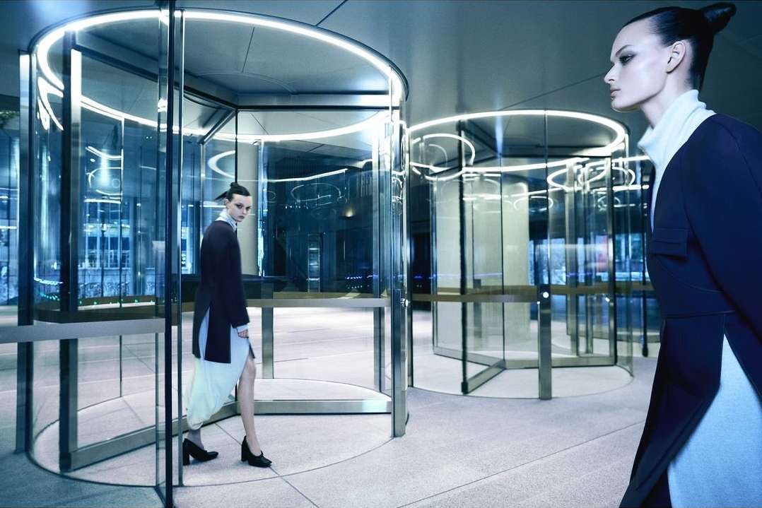 Fashion shot of two women in dark coats walking through modern revolving doors with illuminated frames in an office building lobby.