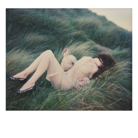 Woman lying down in tall grassy dunes wearing a sheer dress and dark heels, photographed by Emma Summerton.