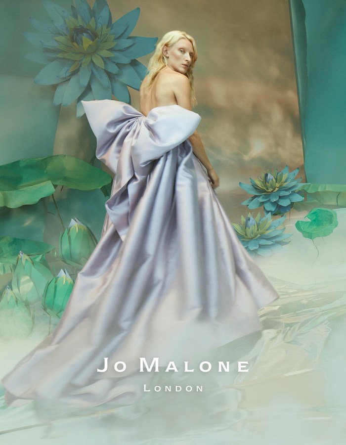 A woman in a lavender gown with a large bow stands in front of light green botanical art for a Jo Malone London advertisement.