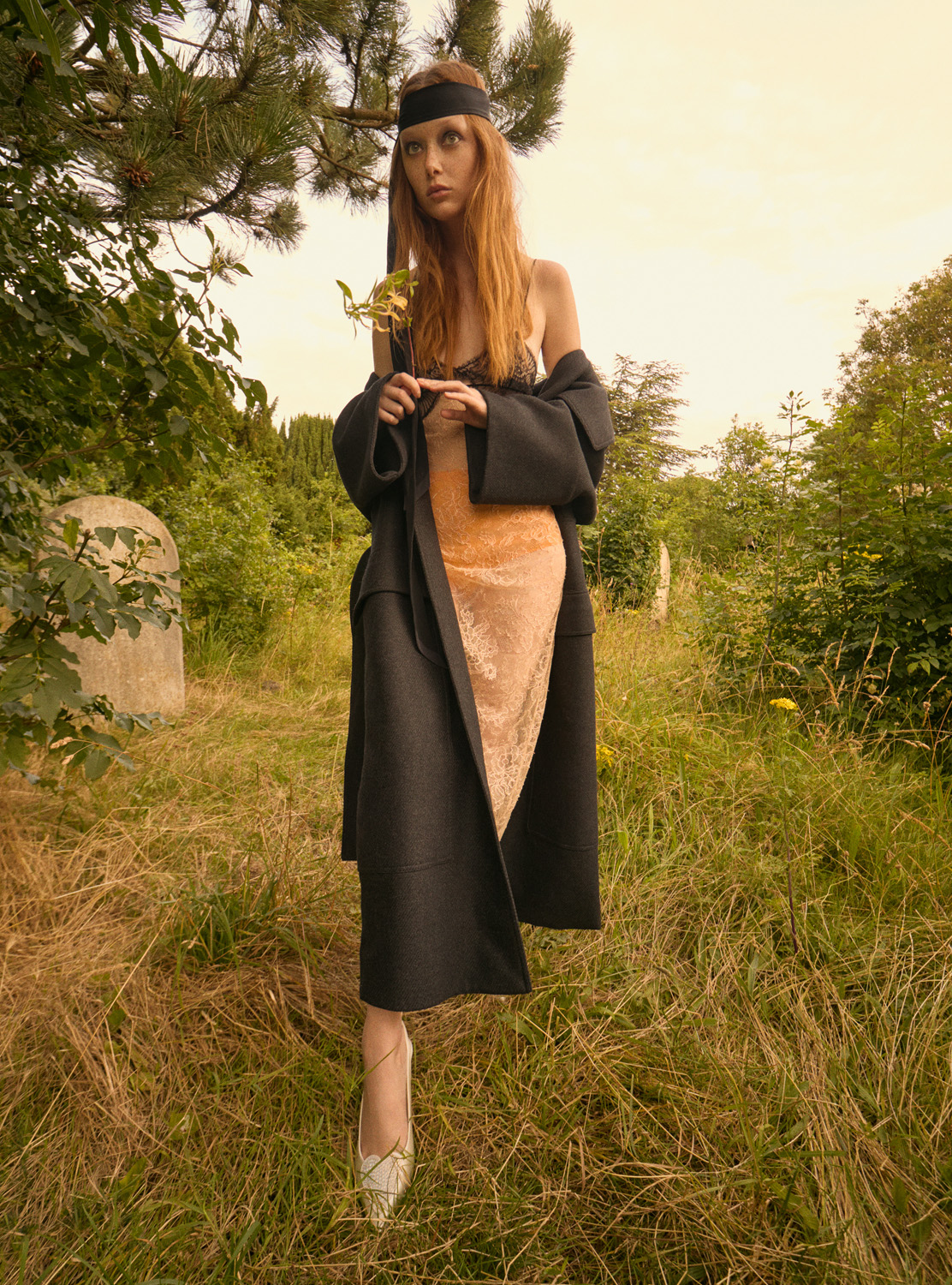 Fashion shot of a redhead model in a black coat and dress, holding a leaf in a grassy field with some tombstone in the background.