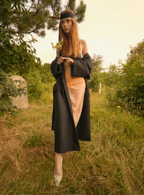 Fashion shot of a redhead model in a black coat and dress, holding a leaf in a grassy field with some tombstone in the background.