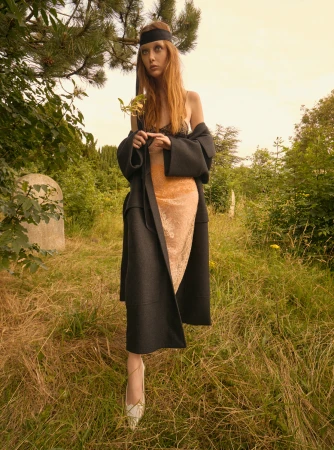 Fashion shot of a redhead model in a black coat and dress, holding a leaf in a grassy field with some tombstone in the background.