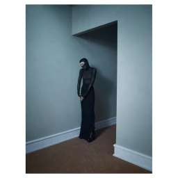 Model wearing a black hooded balaclava and long black gown standing in a dimly lit hallway by Emma Summerton