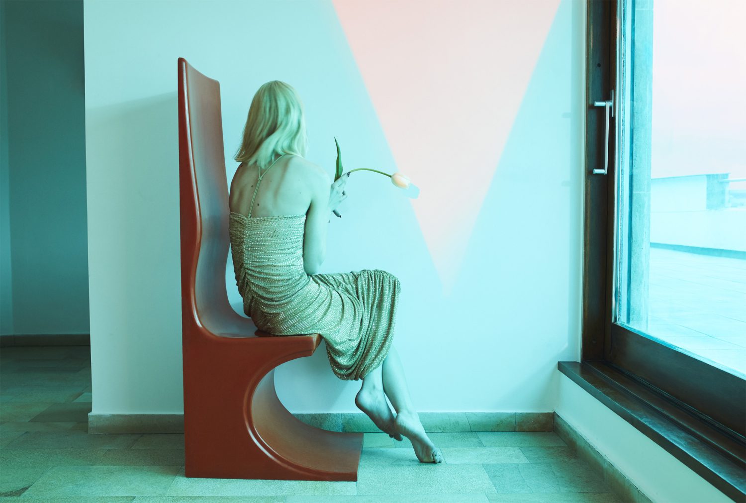 Blonde woman in a textured gold dress seated on a tall brown chair holding a single tulip near a large window by Emma Summerton